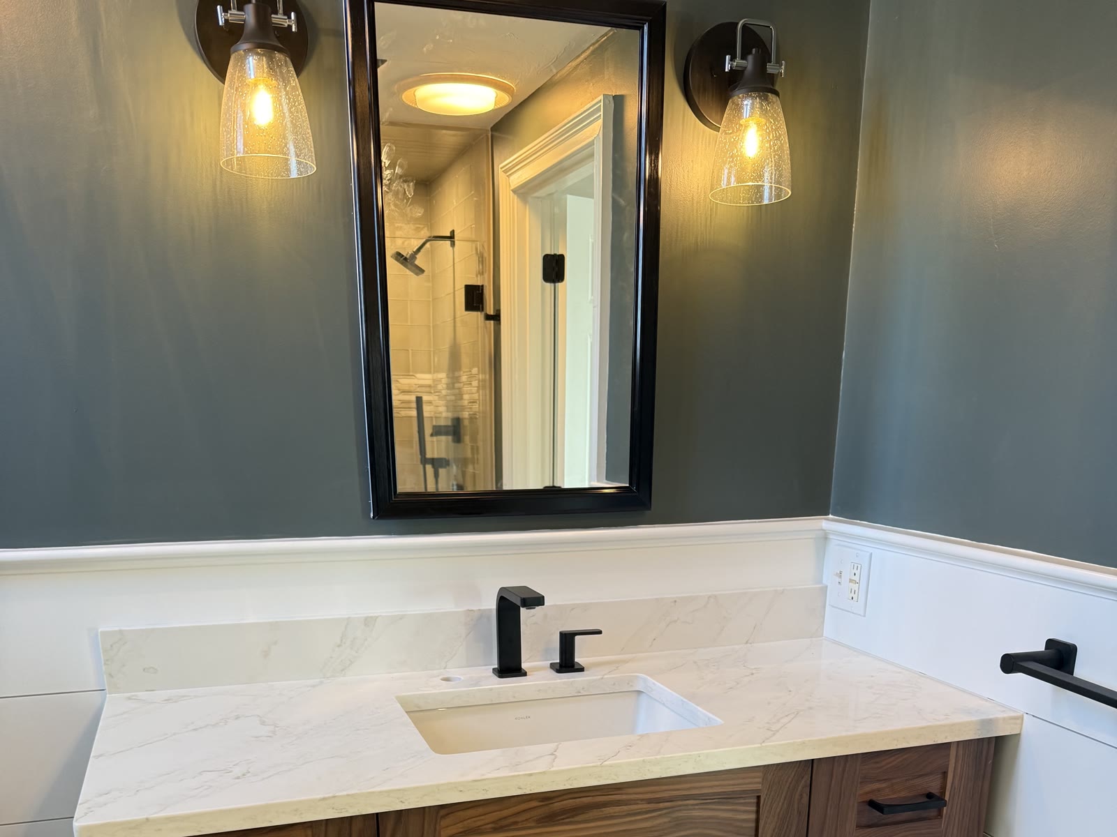 Remodeled bathroom with a walnut vanity, marble countertop, black-framed mirror, and glass wall sconces